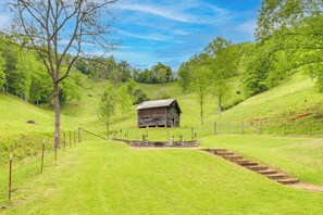 Une cabane en bois rustique, accessible par un sentier de terre, entourée de collines verdoyantes et d’arbres luxuriants.
