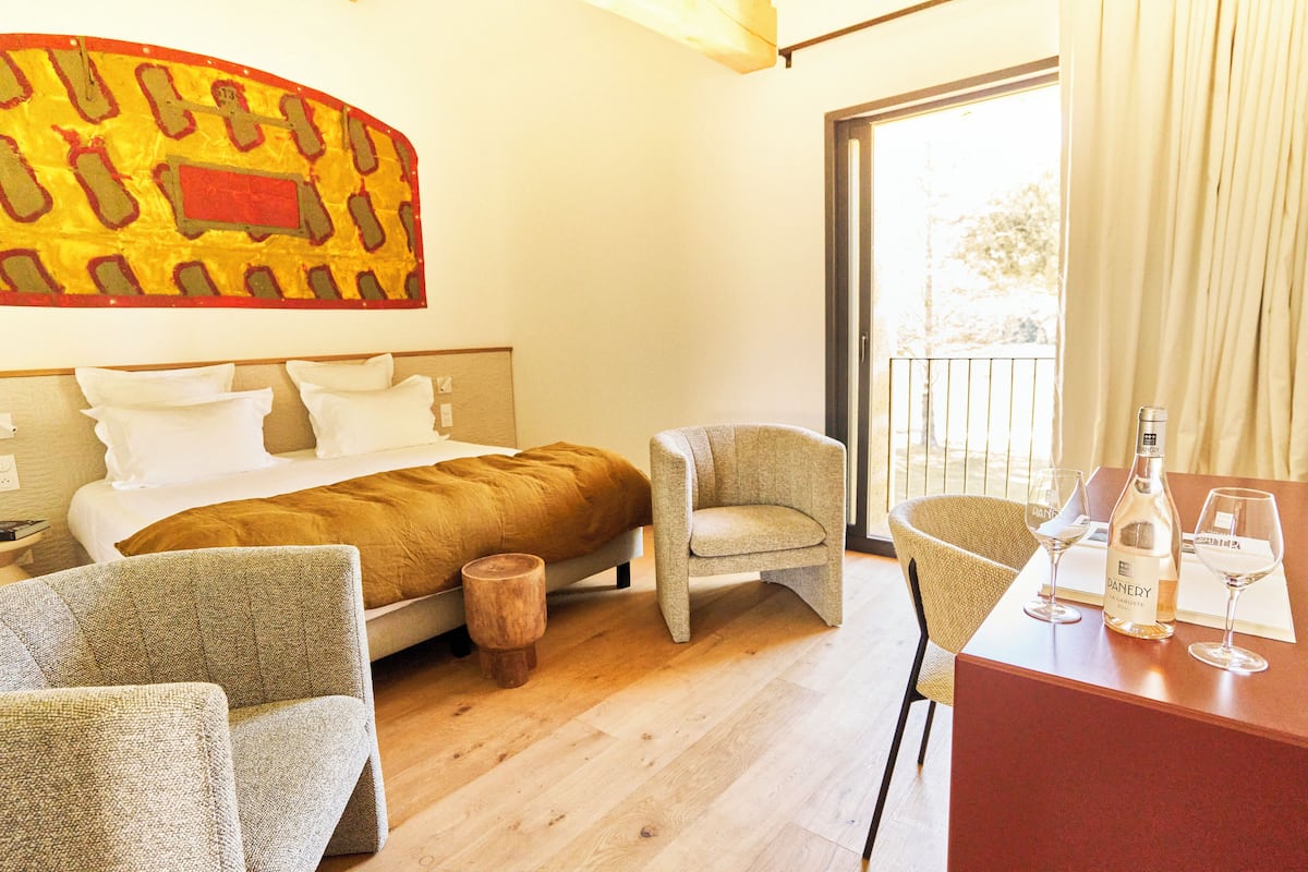 A hotel room with a bed, two armchairs, a wooden stool, a table with a bottle and glasses, and a large window with curtains.