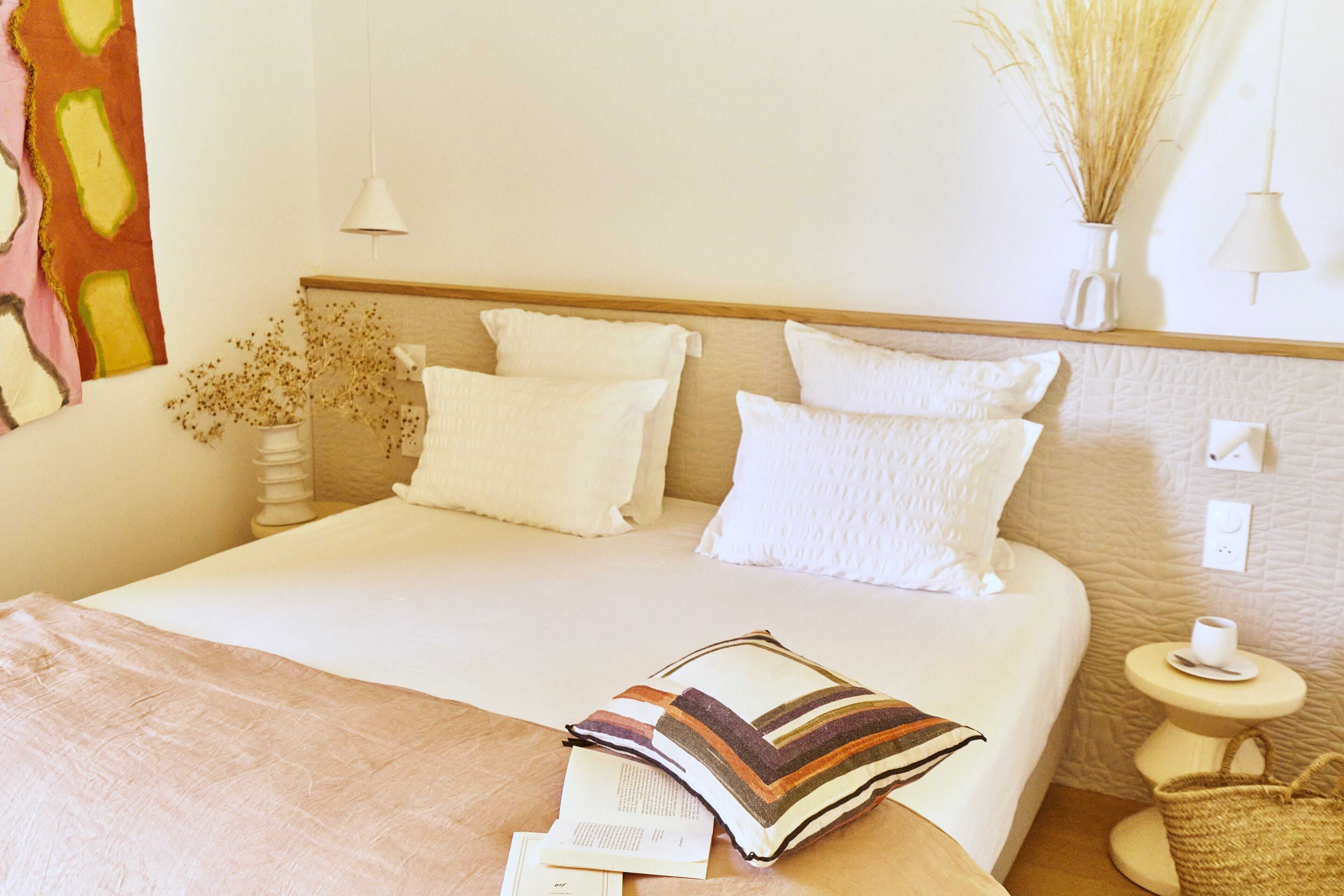 Una cama bien hecha con sábanas blancas, un almohadón a rayas y un libro sobre la cama. Hay una mesita de noche con una taza y su platillo, un florero con plantas secas y una cesta tejida.
