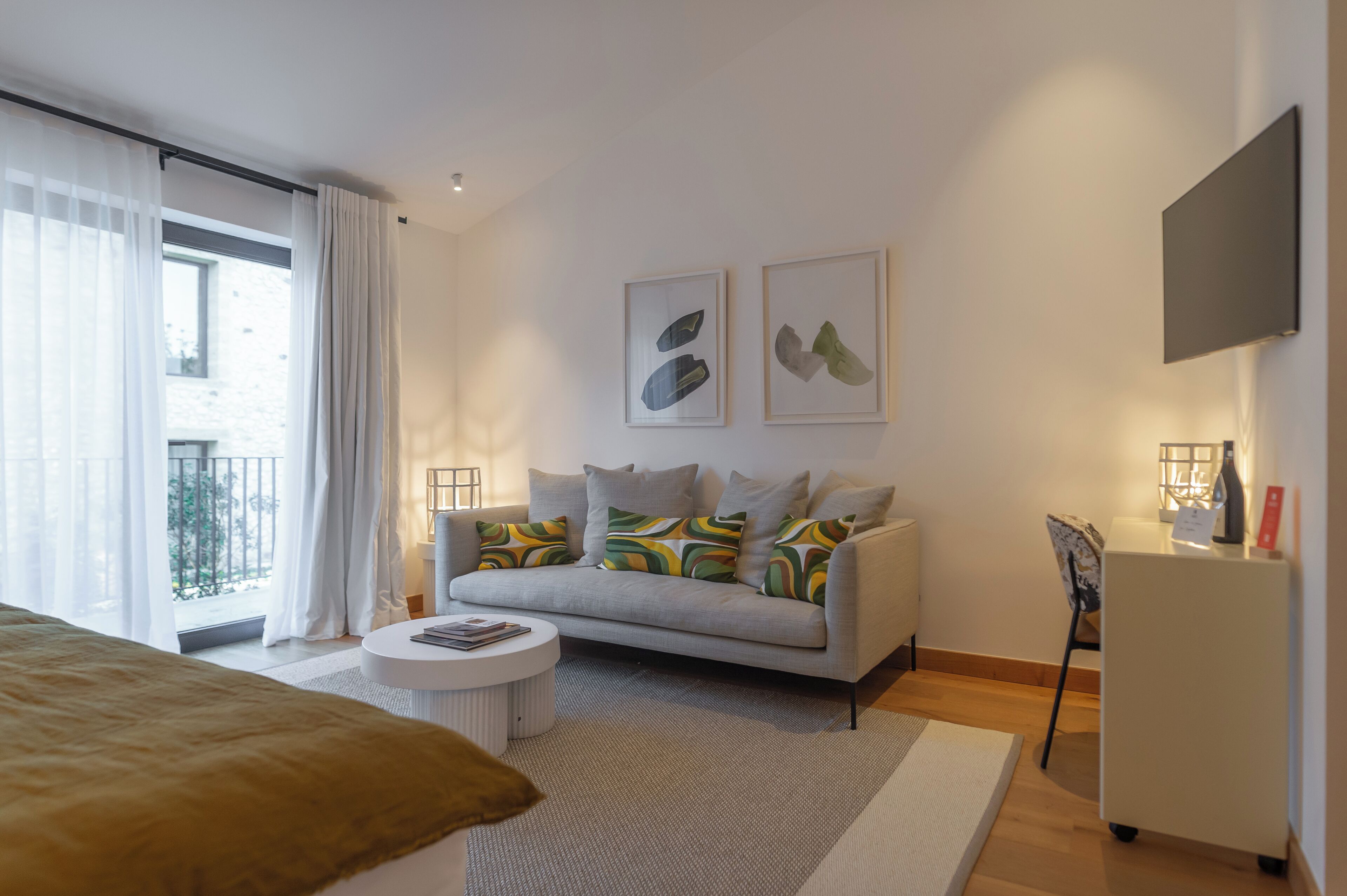 A modern hotel room with a sofa, a small round table, a bed, a TV, and artwork on the wall.