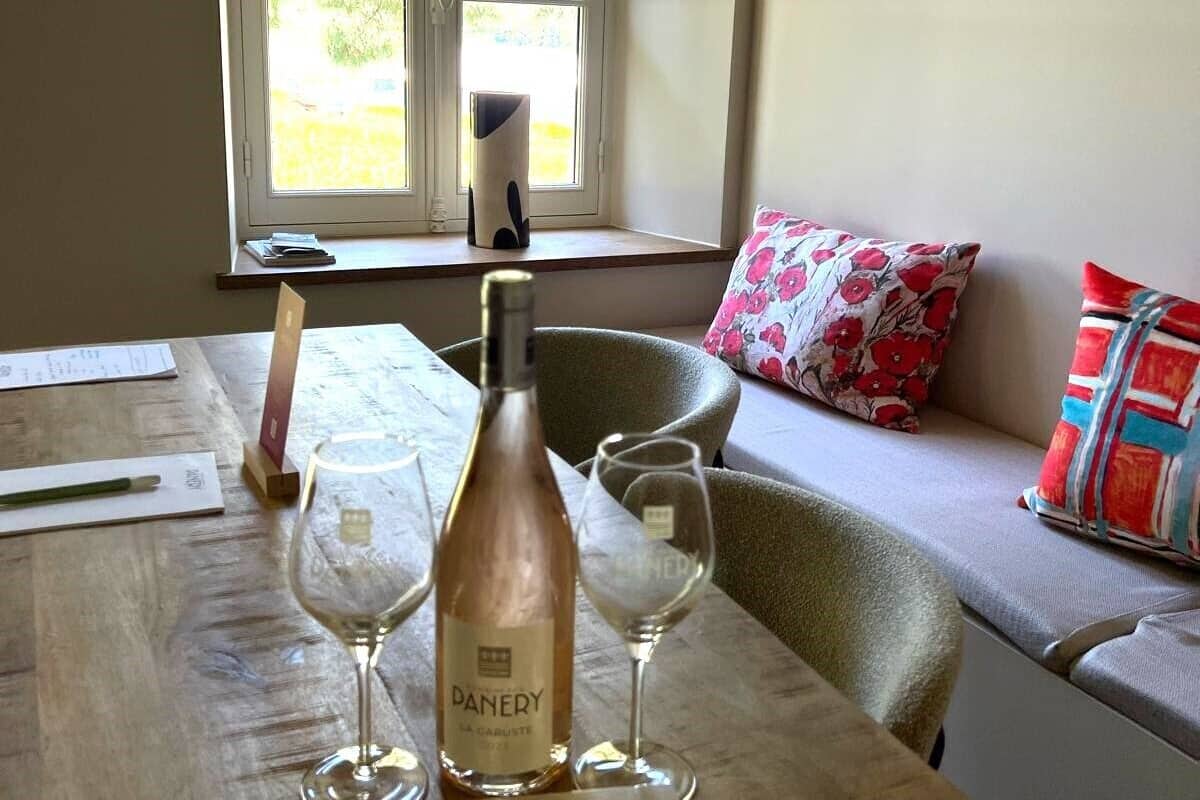 A table with a bottle of wine, two glasses, and a menu card in a room with a window and a sofa with patterned cushions.