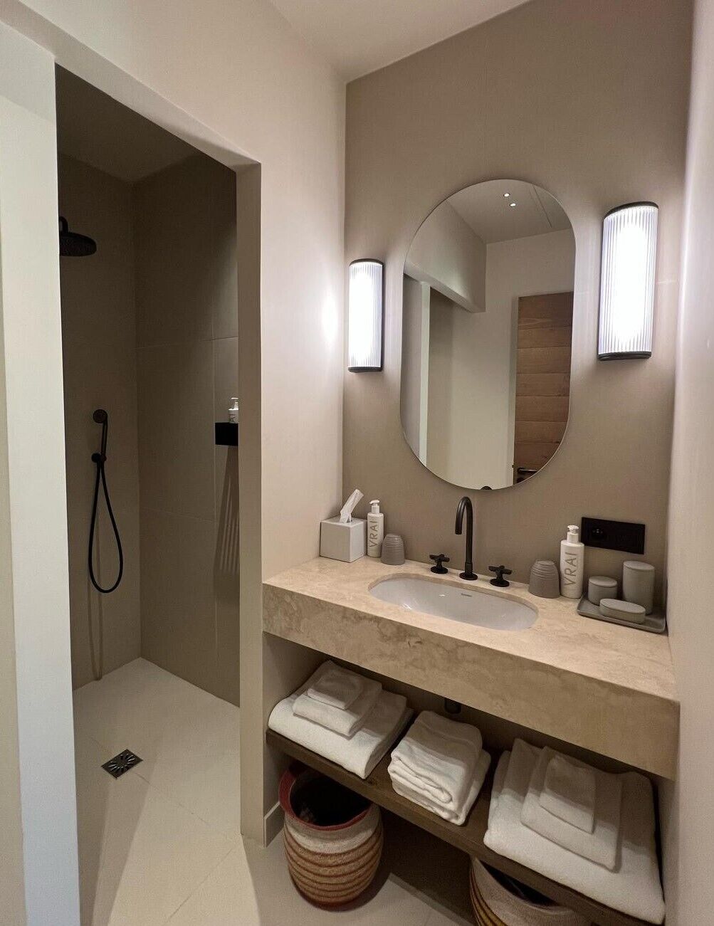 A modern bathroom with a large mirror, a sink, and a shower. There are towels and toiletries on the counter.