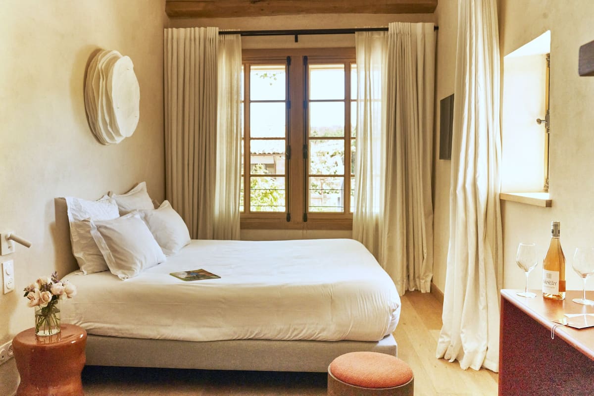 A hotel room with a bed, a bedside table with a bottle and glasses, a vase with flowers, and a window with curtains.