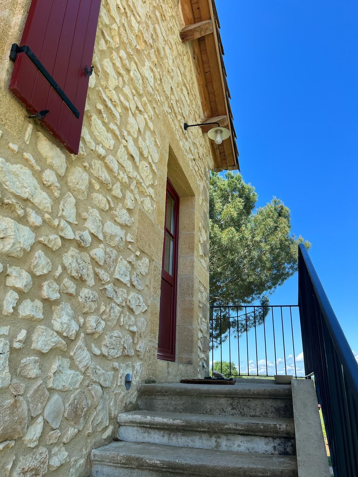 A stone building with a red door and shutter, a wooden staircase, and a metal railing.