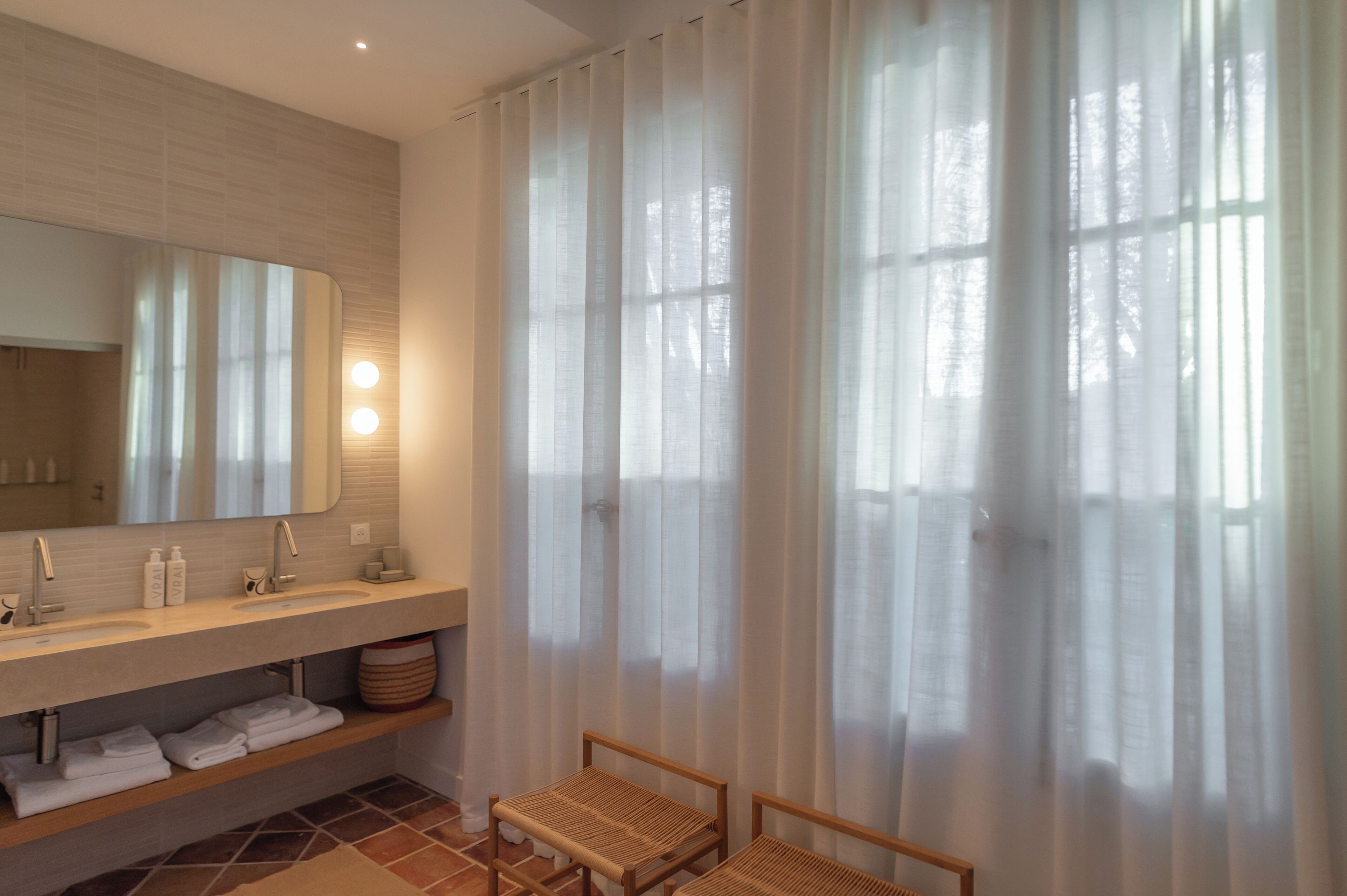 Une salle de bain moderne avec un grand miroir, deux lavabos, un meuble-lavabo et une fenêtre agrémentée de rideaux légers.