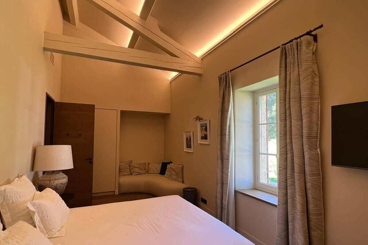 A bedroom with a large bed, a desk, a chair, a lamp, a window with curtains, and a flat-screen TV mounted on the wall.