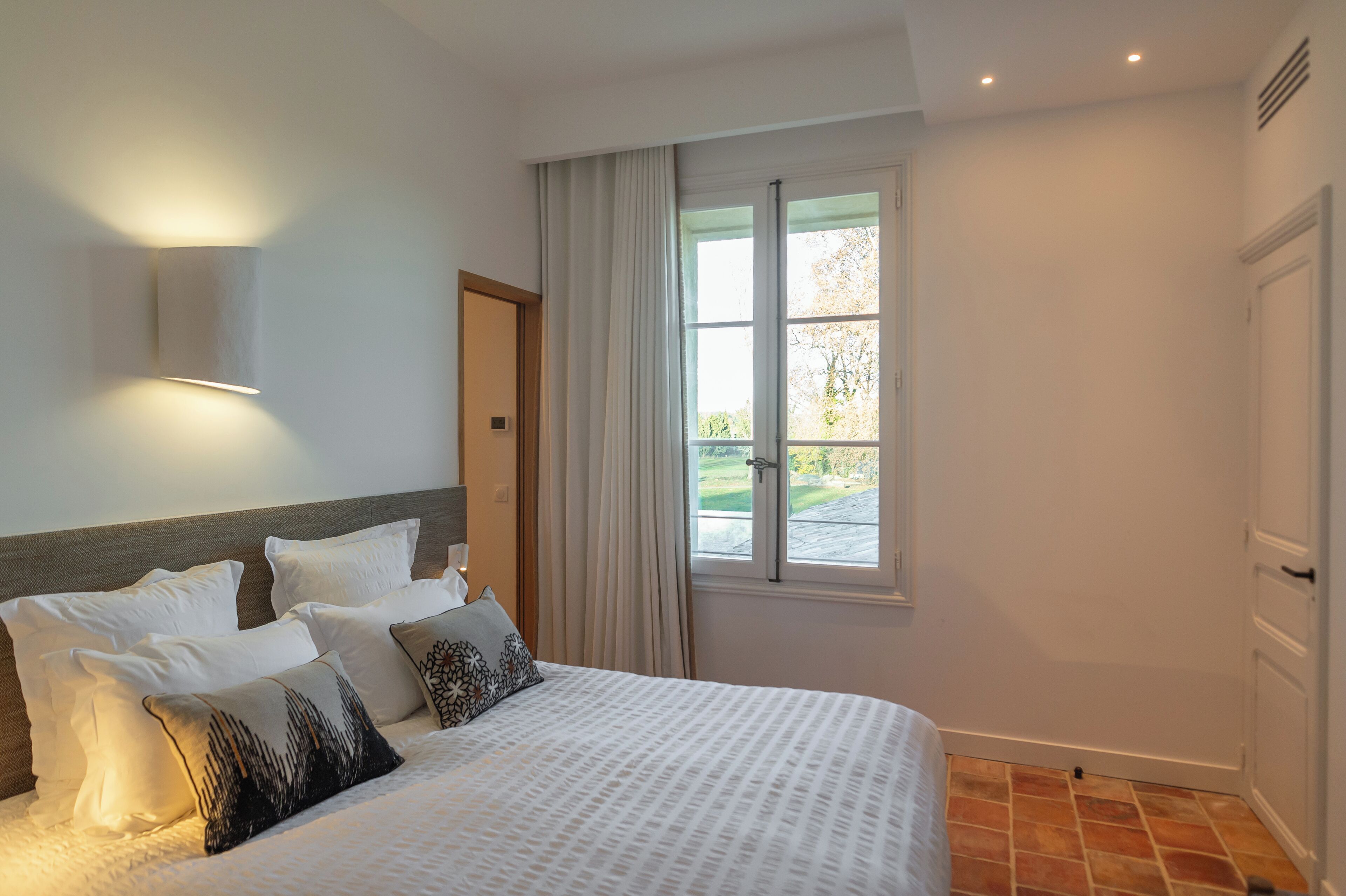 Un dormitorio con cama, ventana con cortinas, luz instalada en la pared y puerta.
