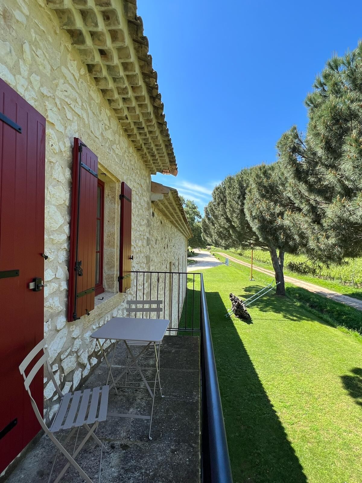 A stone building with red shutters, a small outdoor seating area with a table and chairs, and a well-maintained lawn with trees and a path leading away.