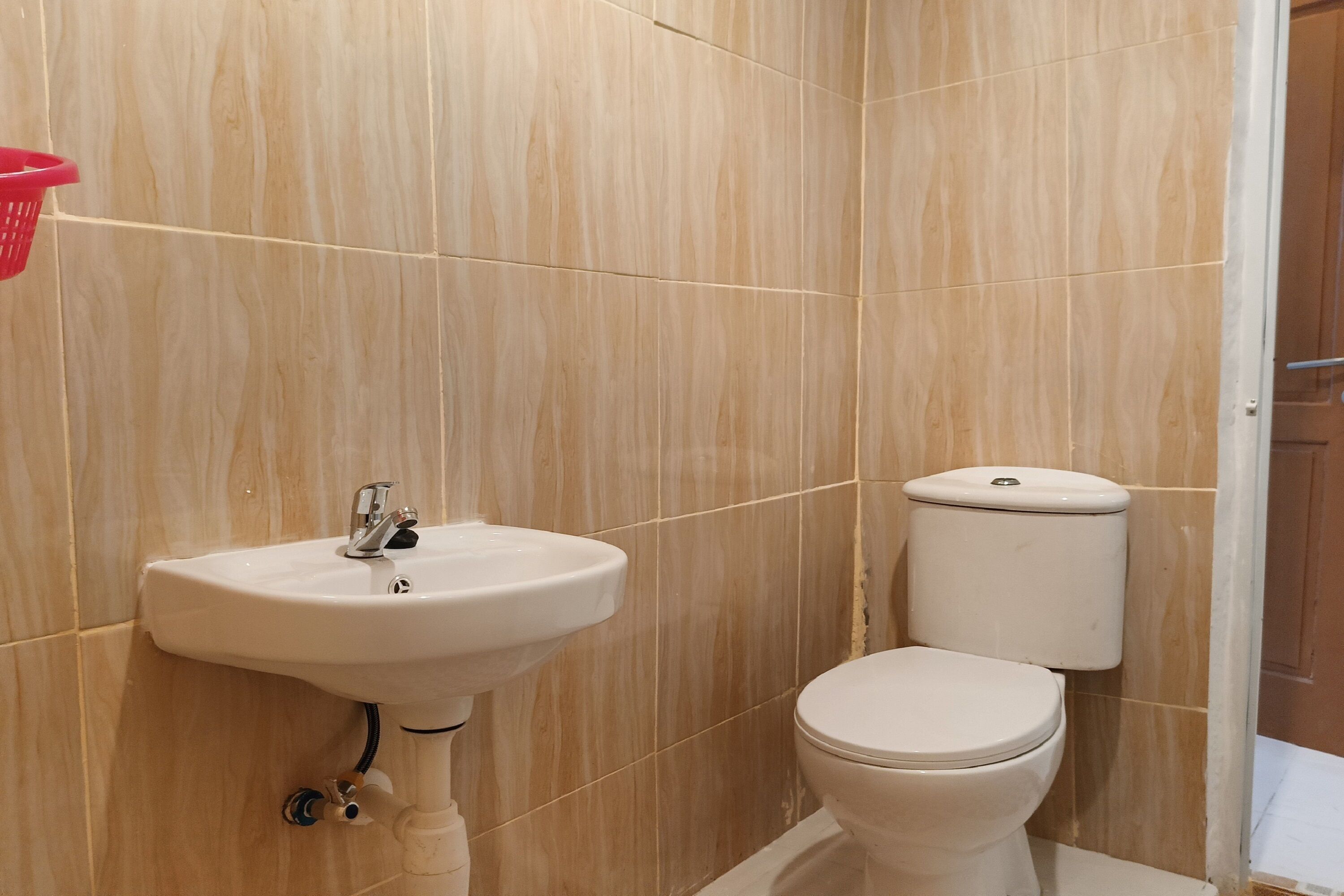 Een badkamer met een witte wastafel en toilet, muren met beige tegels en een zichtbaar deurkozijn.