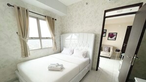A bedroom with a large bed, a window with curtains, a door leading to another room, and a small table with a chair. - OYO 93940 Apartemen Mekarwangi Square By Lina (Bandung)