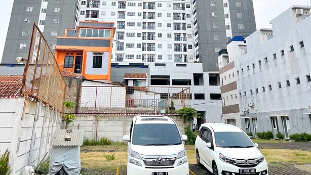 A high-rise building with multiple floors, a parking area with two cars, and a smaller adjacent building with a red roof. - OYO 93940 Apartemen Mekarwangi Square By Lina (Bandung)