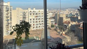 Une pièce dotée d’une grande fenêtre donnant sur un paysage urbain, avec des immeubles résidentiels et un ciel dégagé.