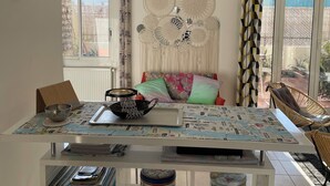 Un coin repas avec une table dressée pour quatre personnes, entourée de chaises. Au-dessus de la table, on peut admirer une décoration murale en macramé, et une fenêtre avec des rideaux offre une vue sur l’extérieur.