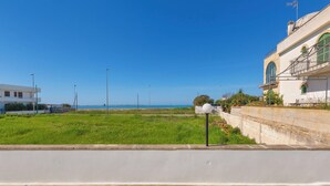 A coastal view with a grassy area, a white building with a balcony, and a clear blue sky. - 3886 Villino Paradiso by Babarhouse (Porto Cesareo)