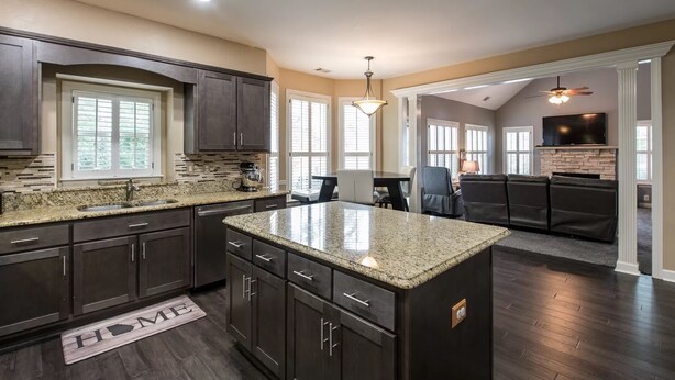 A modern kitchen with dark cabinets, granite countertops, and a central island. There is a dining area with a table and chairs, and a living room with a fireplace and a TV. - NEW Southern Charm ATL 4 Bdr/3 Bath Executive Home (Norcross)