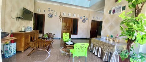 A spacious living area with a high ceiling, a balcony, a dining table with chairs, a kitchen area, and a decorative tree.