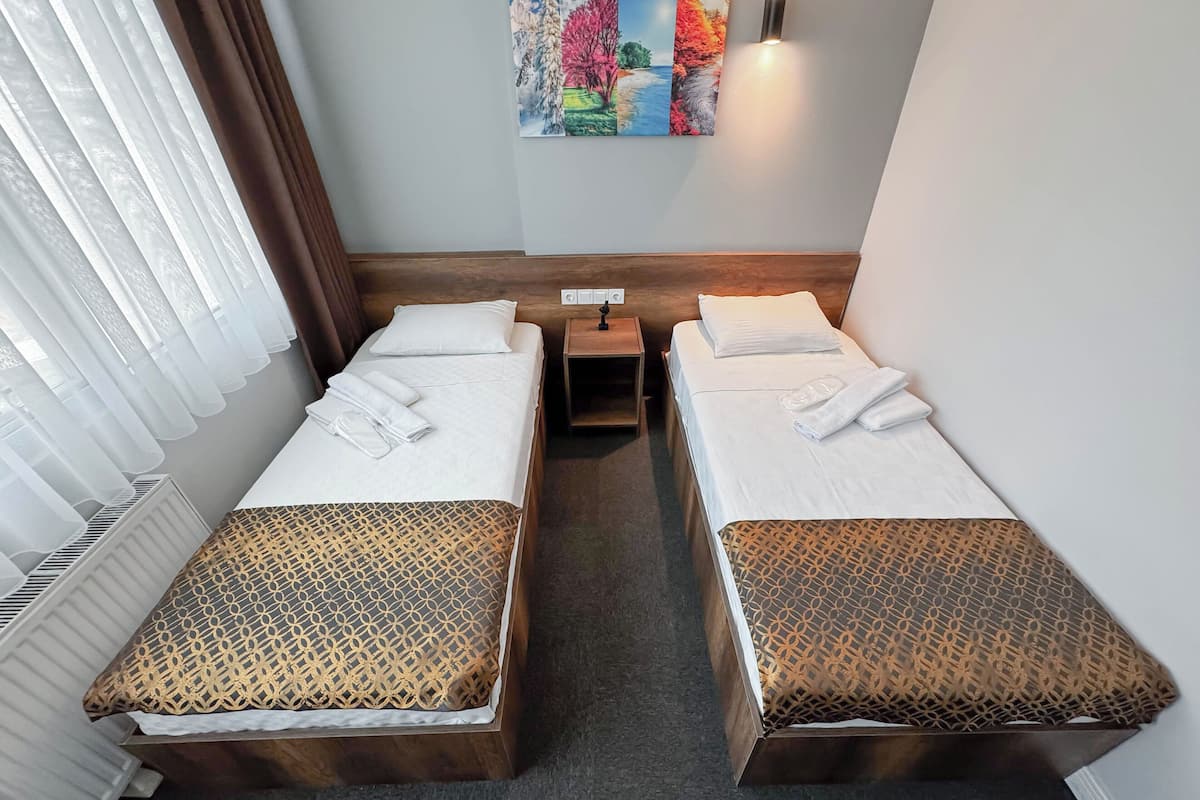 A hotel room with two beds, each with a patterned bedspread and pillows, a nightstand, a painting on the wall, and a window with curtains.