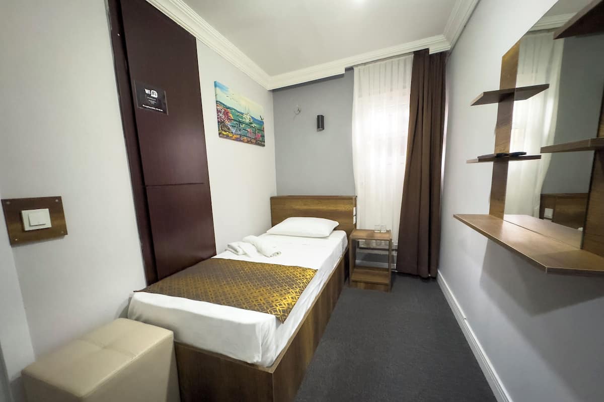 A compact hotel room with a single bed, a small bedside table, a chair, a wardrobe, a mirror, and a painting on the wall.