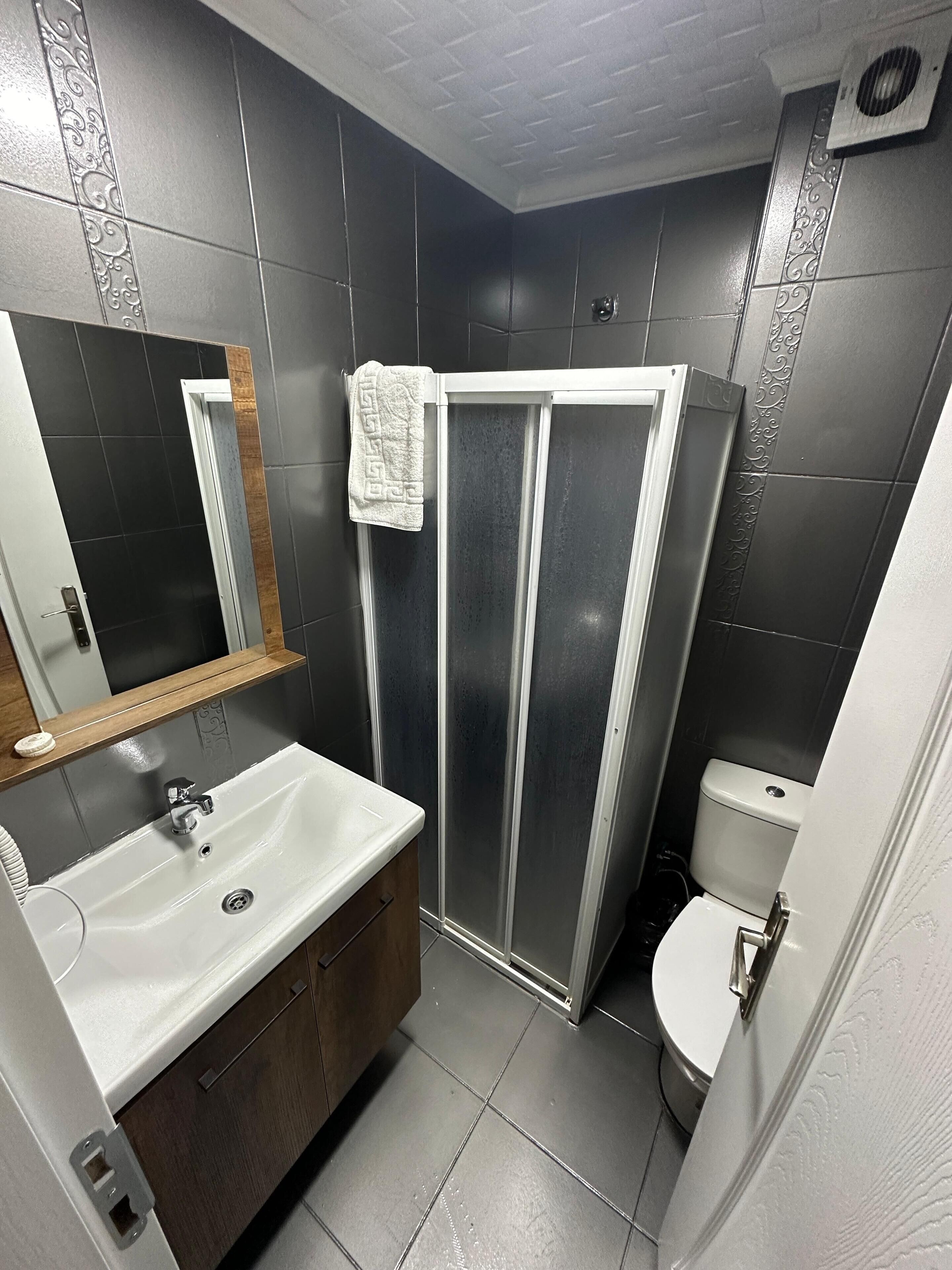 Een moderne badkamer met een wit wastafelmeubel, donker betegelde wanden, een douchecabine en een toilet.