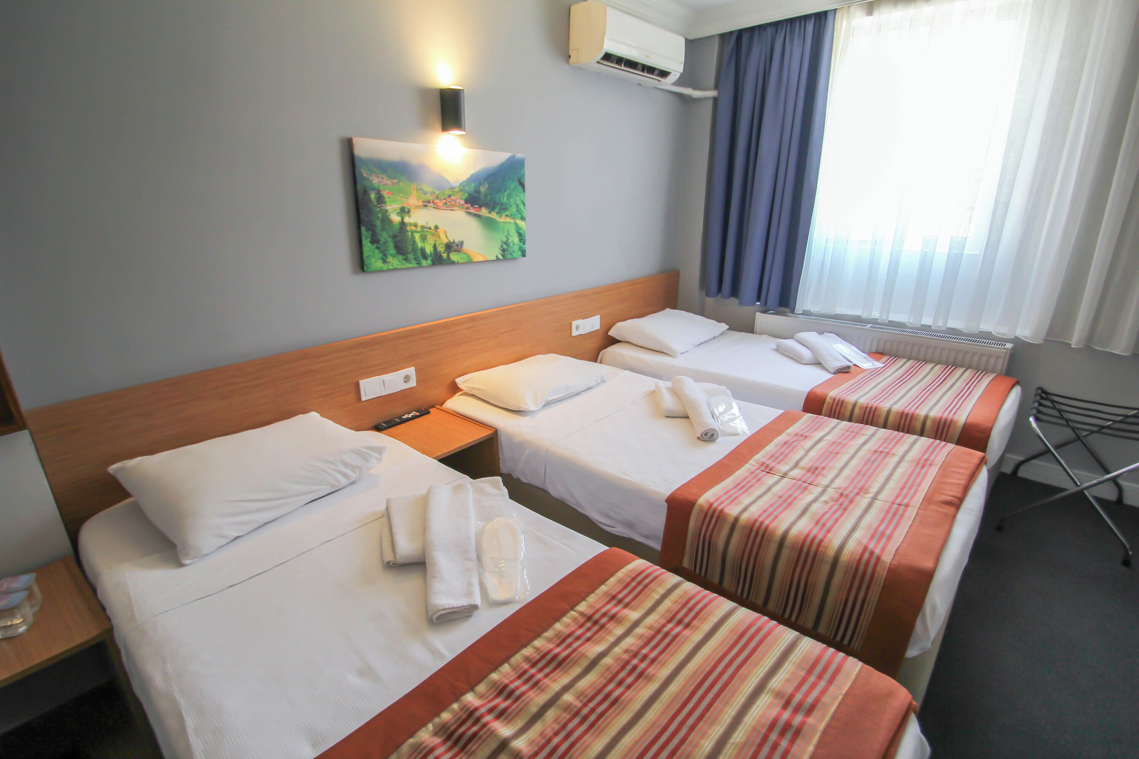 A hotel room with three single beds, each with a white pillow and a neatly folded towel. There is a painting on the wall above the beds, a window with curtains, and an air conditioning unit mounted on the wall.