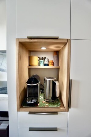 A compact kitchen area with a built-in coffee maker and a kettle, a small shelf with kitchenware, and a drawer below.