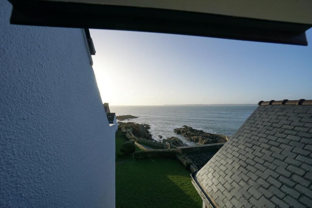 Une vue sur la côte, prise depuis un bâtiment doté d’un toit en tuiles et surplombant un espace verdoyant bordé d’un mur de pierres.