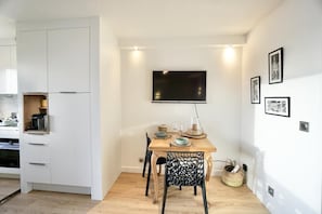 A modern kitchen with white cabinetry, a dining table with chairs, a flat-screen TV mounted above the table, and a small side table with a basket.