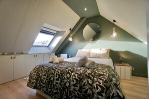A bedroom with a sloped ceiling, a bed with a leaf-patterned bedspread, a round mirror, and a small bedside table.