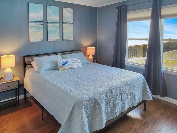 Una cama bien tendida con una colcha azul claro y almohadas, una lámpara de noche y una obra enmarcada sobre la cama.