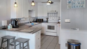 Une cuisine avec des meubles blancs, une cuisinière, un four à micro-ondes et une machine à café. On y trouve deux tabourets de bar et un panneau indiquant : « Bienvenue à la maison de plage ».
