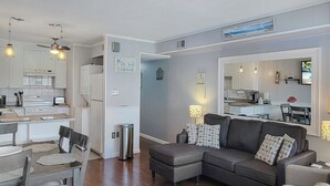 Un salon moderne avec un canapé d’angle gris, un coin repas avec une table et des chaises, et une cuisine avec des meubles blancs et une cuisinière.