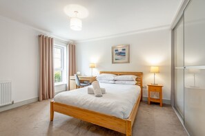 A bedroom with a wooden bed, a desk, a chair, a lamp, and a window with curtains. - Kingsbarns House (No 1) (St Andrews)