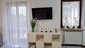 Un salón con un televisor montado en la pared, una consola blanca con un florero y una ventana con cortinas ligeras.