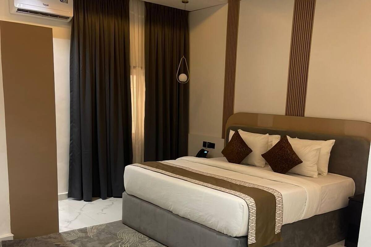 A modern hotel room with a large bed, a bedside table, a wall-mounted air conditioner, and a patterned rug.