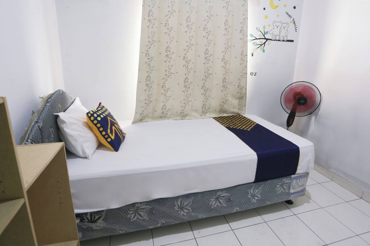 A single bed with a white mattress, a blue blanket, and a floral-patterned headboard. A small fan is on the floor beside the bed.