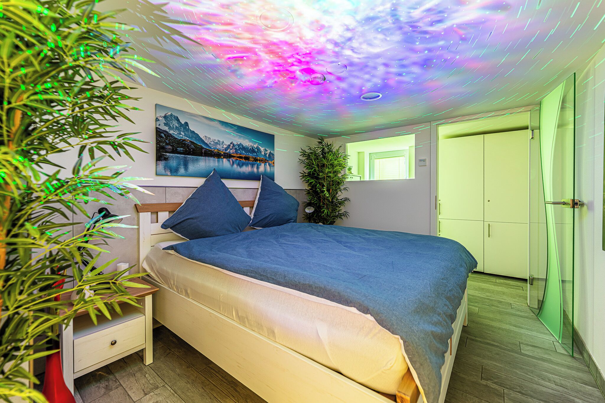 A bedroom with a bed, a nightstand, a plant, a painting, and a door.