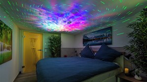 Een slaapkamer met een bed, nachtkastje, lamp, plant en een schilderij aan de muur.