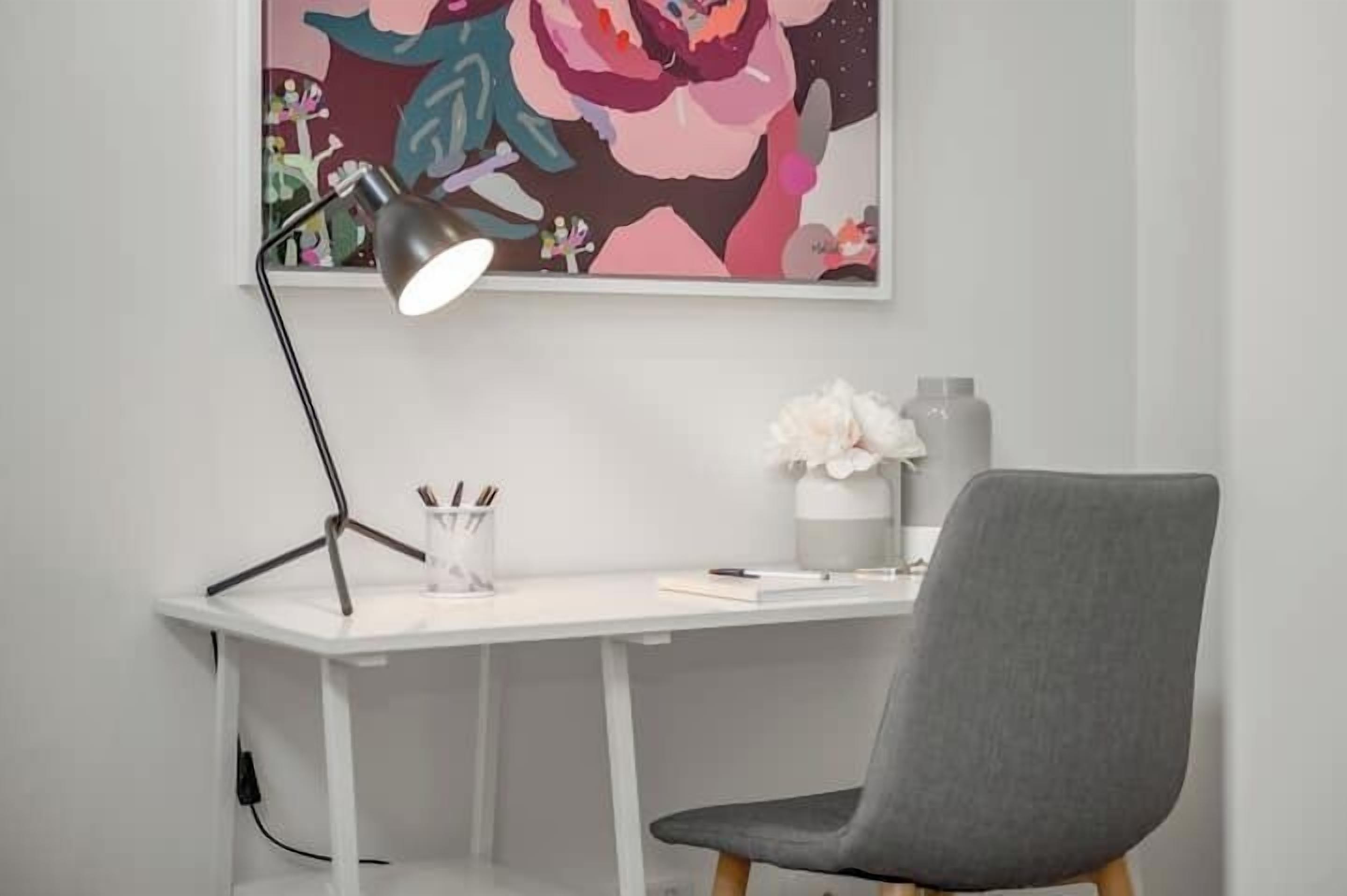 Un bureau avec une lampe, une chaise, un vase de fleurs et un tableau coloré accroché au mur.