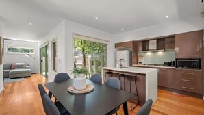 A modern kitchen with a dining area, wooden flooring, and a view of greenery outside.