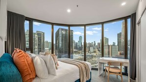 Un dormitorio moderno con una cama curvada, un escritorio y vistas a un horizonte urbano a través de amplias ventanas.