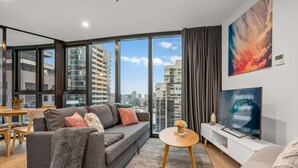 Una sala de estar moderna con un sofá, una mesa de centro, televisor y ventanas amplias con vista a la ciudad.