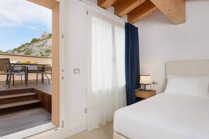 A hotel room with a bed, a nightstand, a lamp, a window with curtains, a balcony with a table and chairs, and a view of a rocky landscape. - Sunset deluxe - Portopiccolo Apartments (Duino-Aurisina)