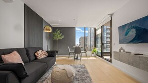A modern living room with a black sofa, a grey rug, a dining area with a table and chairs, and a large window offering a city view. - Executive Designer Dreaming Home with All you need (Moonee Ponds)