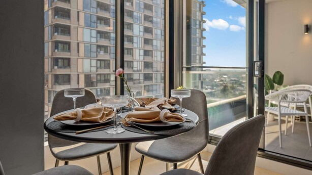 A dining area with a table set for two, a view of a high-rise building, and a balcony with a chair. - Executive Designer Dreaming Home with All you need (Moonee Ponds)