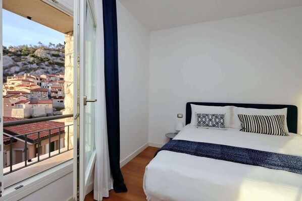 A hotel room with a balcony, a bed with white bedding and patterned pillows, a small bedside table, and a view of a hillside town.