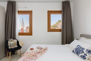 Ein Schlafzimmer mit einem Bett, zwei mit Vorhängen versehenen Fenstern, einem Stuhl und einer Tasche sowie Blick auf Gebäude und Hügel.