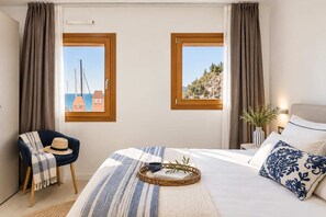 A hotel room with a bed, a chair, a window with a view of a coastal landscape, and a bedside table with a lamp and a vase.