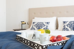 A neatly made bed with a tray of breakfast items including fruits and cups, a bedside lamp, and patterned pillows.