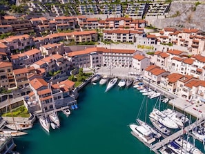 Ein Luftbild einer Küstenstadt mit einem Yachthafen, das mehrstöckige Gebäude mit roten Ziegeldächern, einen zentralen Platz und Boote im Hafen zeigt.