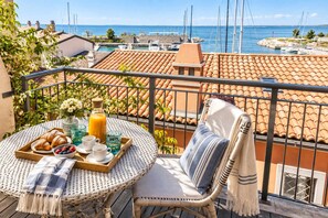 A balcony with a wicker table set for breakfast, overlooking a marina with boats and a clear sky.